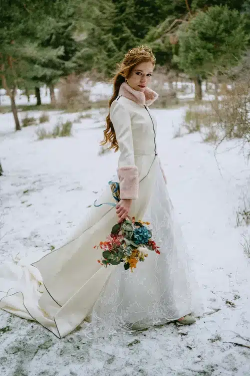 la novia con el abrigo en movimiento sujetando el ramo y posando en la nieve